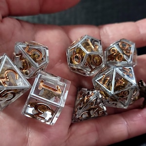 May include: A set of seven polyhedral dice made of clear resin with gold-colored numbers. Each die contains small, textured, gold-toned metallic inclusions suspended within the transparent material, visible through the faceted surfaces.