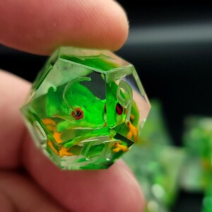Peut inclure: Un dé transparent à multiples facettes avec une grenouille verte vibrante à l'intérieur. La grenouille a des yeux rouges et des pieds orange, sur un fond clair. D'autres dés sont visibles en arrière-plan. Le dé est tenu entre les doigts.