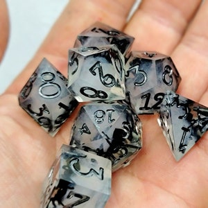 May include: A set of seven clear dice with black ink, featuring a variety of shapes and numbers. The dice are translucent and have a cloudy, marbled appearance.