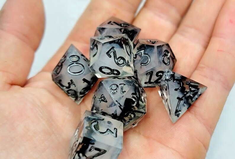 May include: A set of seven clear dice with black ink, featuring a variety of shapes and numbers. The dice are translucent and have a cloudy, marbled appearance.