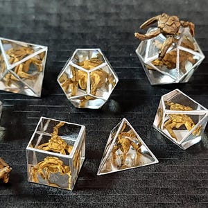 May include: A set of seven polyhedral dice made of clear resin, each containing a small, detailed brown crab figure inside. The collection includes various geometric shapes such as cubes, pyramids, and multi-faceted dice, all featuring sharp edges.