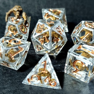 May include: A set of seven polyhedral dice made of clear resin with copper-colored numbers. Each die contains a small, brown, skeletal-like structure suspended inside the transparent material. The set includes various geometric shapes for gaming.