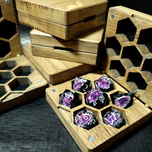 Könnte beinhalten: Würfelboxen aus Holz mit Wabenmuster. Die Boxen sind aus hellbraunem Holz gefertigt und enthalten einen Satz lila und schwarzer Würfel. Die Boxen sind geöffnet und geschlossen, wobei die Würfel im Inneren zu sehen sind.