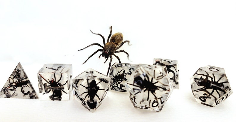 May include: A set of six clear dice with black numbers and a spider encased in each die. A larger spider is in the background.