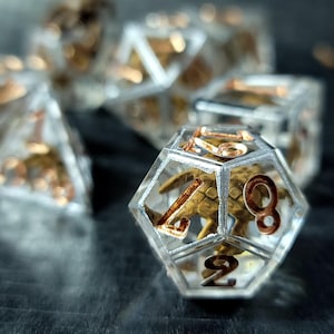 May include: A set of polyhedral dice made of clear resin with sharp edges. Each die contains a small, gold-colored metallic dragon figure suspended inside. The numbers on the faces are engraved and filled with a metallic copper-colored pigment.