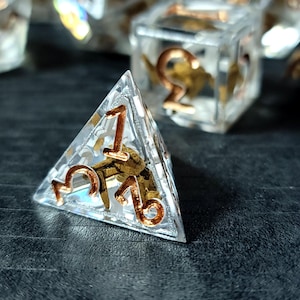 May include: A four-sided triangular die made of clear resin with metallic copper-colored numbers embedded on the surface. The die features a small, gold-toned metallic scorpion figure suspended inside the transparent material, visible through the clear faces.