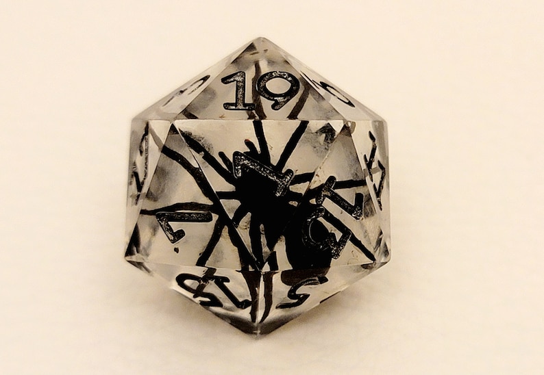 May include: A clear, multi-sided dice with black numbers and a spider design. The dice is a d20, with the number 19 visible.