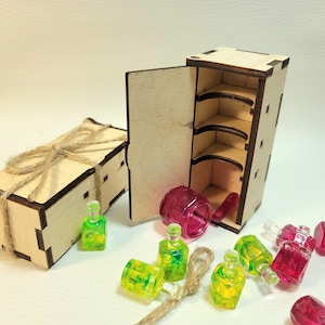 May include: A wooden box with a hinged door and shelves, open to reveal a pink plastic container. The box is made of light wood with dark brown accents. There are several small green and pink glass bottles on a white surface.