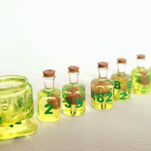 May include: A set of seven green and yellow glass bottles with cork tops. Each bottle contains a small, green, numbered dice. The dice are numbered 1 through 7.