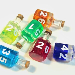 May include: Six colourful glass dreidels with white numbers on each side. The dreidels are filled with liquid and have a stopper on top. The dreidels are arranged in a circle on a white surface.