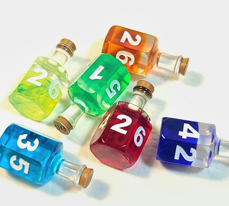 May include: Six colourful glass dreidels with white numbers on each side. The dreidels are filled with liquid and have a stopper on top. The dreidels are arranged in a circle on a white surface.