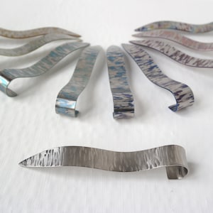 May include: A set of nine silver bookmark with a wavy design and a loop at the end. The bookmarks have a brushed metal finish and are decorated with a variety of patterns.