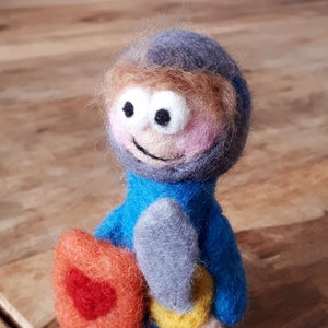 May include: A handmade felt knight figurine. The knight is wearing a blue tunic and helmet, with a grey sword and orange shield with a red heart. The knight has a smiling face with large eyes.