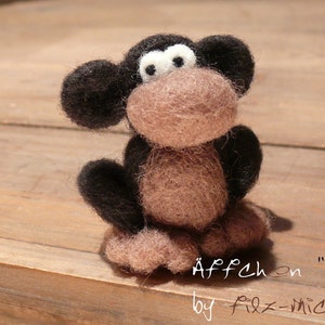 Può includere: Un giocattolo di scimmia di feltro marrone e nero con occhi bianchi. Il giocattolo è seduto su una superficie di legno. Il testo "Äffchen "Bobo" by Filz-Michel" è scritto sotto il giocattolo.