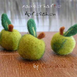 May include: Three green felt apples with brown stems and green leaves. The apples are arranged on a wooden surface. The text "zauberhafte Apfelchen" and "by Alex-Michel" are written on the surface.
