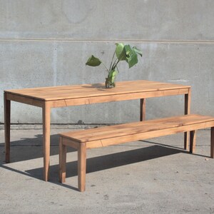 May include: A light brown wooden dining table with a matching bench. The table has a rectangular top and four legs. A clear glass vase with green leaves sits on the table. The bench is similar in design to the table.