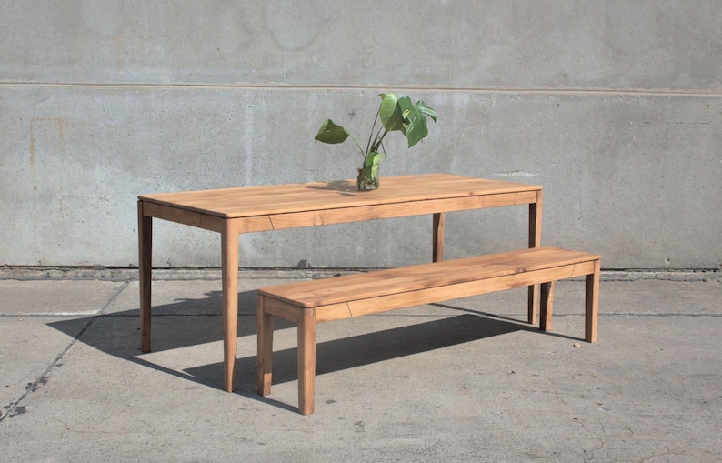 May include: A light brown wooden dining table with a matching bench. The table has a rectangular top and four legs. A clear glass vase with green leaves sits on the table. The bench is similar in design to the table.