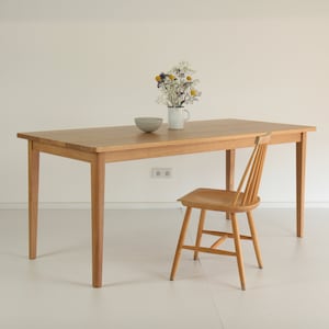 Könnte beinhalten: Ein heller Holz-Esstisch mit einem Stuhl. Der Tisch hat ein schlichtes Design mit geraden Beinen und einer rechteckigen Tischplatte. Eine kleine weiße Schüssel und eine weiße Tasse mit einem Strauß weißer und gelber Blumen stehen auf dem Tisch.