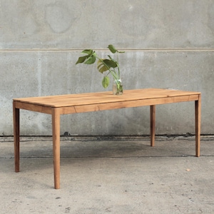 May include: A rectangular wooden table with four legs. The table is light brown and has a natural wood grain. A small glass vase with green leaves sits on the table's surface. The table is set against a gray concrete wall.
