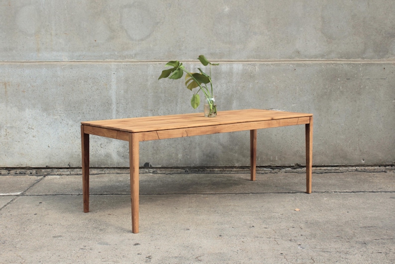 May include: A rectangular wooden table with four legs. The table is light brown and has a natural wood grain. A small glass vase with green leaves sits on the table's surface. The table is set against a gray concrete wall.