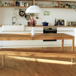 May include: A wooden dining table and bench set in a bright kitchen. The table and bench are made of light-colored wood. The kitchen has white cabinets, a white sink, and a stainless steel oven. A white pendant light hangs above the table.