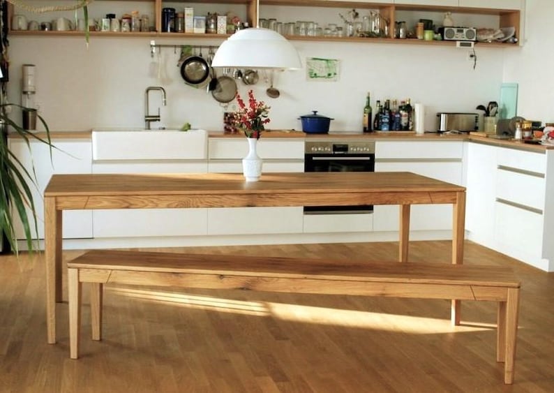 May include: A wooden dining table and bench set in a bright kitchen. The table and bench are made of light-colored wood. The kitchen has white cabinets, a white sink, and a stainless steel oven. A white pendant light hangs above the table.