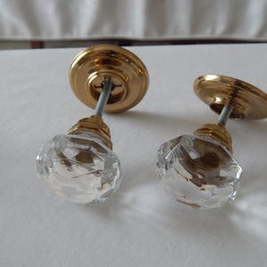 May include: Two decorative door knobs. Each knob features a faceted, clear crystal glass handle and a gold-colored metal base and backplate. The knobs are designed for installation on a door.