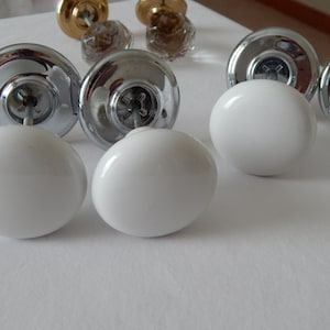 May include: A selection of door knobs in various styles. Some are chrome with white, rounded porcelain knobs, while others are gold-toned and clear glass. The knobs are displayed on a white surface, highlighting their different designs and finishes.