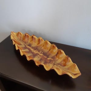 May include: A leaf-shaped ceramic dish with a ruffled edge. The dish has a blend of brown and yellow tones, creating a natural aesthetic. The elongated design is suitable for serving or display. The dish is placed on a dark brown surface.