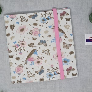 Könnte beinhalten: Ein weißes Stoffnotizbuch mit einem rosafarbenen Blumenmuster und einem rosafarbenen Bandverschluss. Das Notizbuch hat einen braunen Hintergrund mit rosa und blauen Blumen, Schmetterlingen und Vögeln. Der Text "made by Caliadu" ist auf einer weißen Karte mit einem rosa Herzen gedruckt.