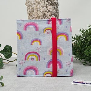Könnte beinhalten: Eine hellblaue Stofftasche mit einem Regenbogenmuster und einem roten Gummizugverschluss. Die Tasche ist mit rosa, gelben und violetten Regenbögen, weißen Sternen und rosa Herzen verziert.