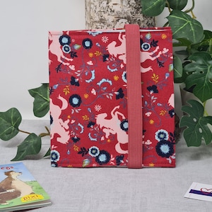 Könnte beinhalten: Ein roter Buchumschlag mit einem rosafarbenen Blumenmuster und einem rosafarbenen Gummibandverschluss. Das Muster zeigt rosafarbene Pferde und Blumen. Der Buchumschlag ist aus Stoff.