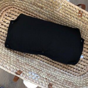 Könnte beinhalten: Ein schwarzes Stoff-Babynest mit Knopfverschlüssen, das in einem geflochtenen Weidenkorb liegt.