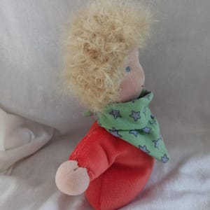 May include: A handmade doll with blonde curly hair, wearing a red shirt and a green scarf with blue stars.