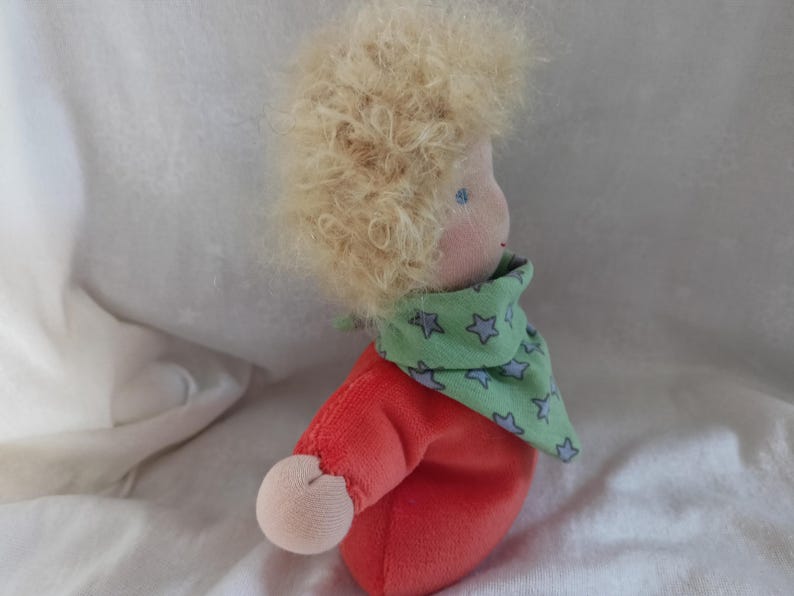 May include: A handmade doll with blonde curly hair, wearing a red shirt and a green scarf with blue stars.