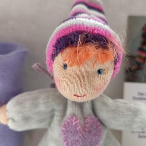 Könnte beinhalten: Handgemachte Puppe mit Stoffkörper, trägt ein graues Outfit mit einem lila Herz und einem gestreiften Hut in Rosa, Lila und Weiß. Die Puppe hat orangefarbene Haare und blaue Augen. Ein lila Filzstiefel ist im Hintergrund.