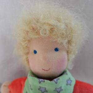 May include: A handmade doll with blonde curly hair, blue eyes, and a green and white star patterned scarf. The doll is wearing a red shirt.