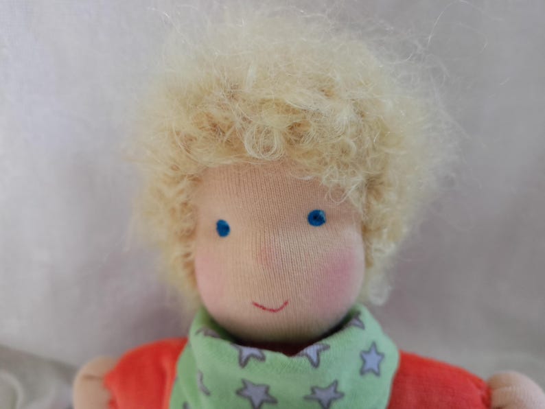 May include: A handmade doll with blonde curly hair, blue eyes, and a green and white star patterned scarf. The doll is wearing a red shirt.