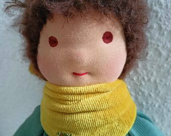May include: A handmade doll with brown curly hair, a light pink face, red embroidered eyes, and a red stitched smile. The doll is wearing a green shirt and a yellow scarf with a red embroidered flower.