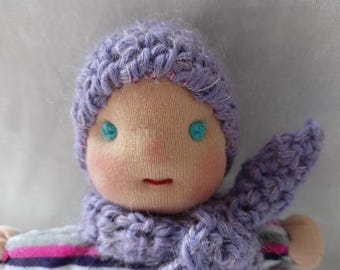 May include: A handmade doll with a light skin tone, wearing a lavender knitted hat and scarf. The doll has turquoise eyes, a small red mouth, and rosy cheeks. The doll's clothing includes a striped shirt with pink, white, and purple stripes.