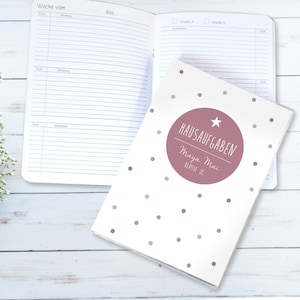 Pode incluir: Um caderno branco com uma capa de bolinhas rosa. A capa tem um círculo rosa com o texto "HAUSAUFGABEN" e o nome "Maya Mai" em um círculo branco. O caderno está sobre uma superfície de madeira branca.
