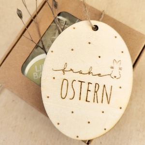 Może przedstawiać: Drewniana ozdoba wielkanocna z napisem "Frohe Ostern" i małym sylwetką królika. Ozdoba jest jasnobrązowa i ma otwór na górze do zawieszenia.