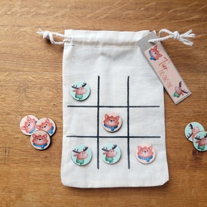 Può includere: Un set di gioco Tris con una borsa di tela con una griglia, pedine di legno con illustrazioni di cervi e volpi e un'etichetta con la scritta "Tier Tic-Tac-Toe". I pezzi del gioco sono sparsi su una superficie di legno.