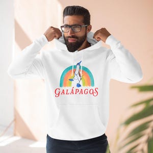 May include: White hooded sweatshirt featuring a colorful rainbow design with a blue-footed booby bird and the word "GALÁPAGOS" in red. The sweatshirt has a front pocket and a drawstring hood.