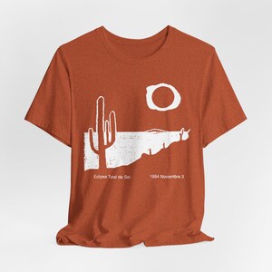 May include: Terracotta-coloured t-shirt with a white desert landscape graphic, including a cactus, sun, and text. The text says "Eclipse Total de Sol" and "1994 Noviembre 3". The shirt has short sleeves and a round neck.