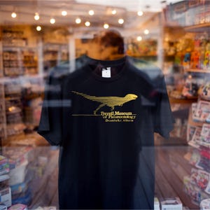 May include: Black t-shirt featuring a gold graphic of a dinosaur and the text "Tyrrell Museum of Palaeontology Drumheller, Alberta." The shirt is displayed in a store window, with reflections of the interior visible.