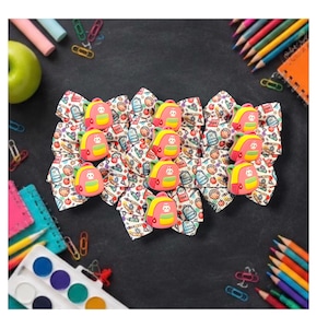 May include: A collection of hair bows featuring a white fabric base with a colorful school-themed pattern of pencils, apples, and books. Each bow is centered with a three-dimensional rubber charm shaped like a pink and yellow backpack.