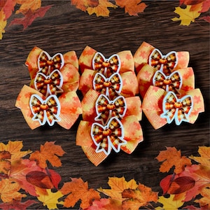 May include: A collection of orange and yellow hair bows with a fall theme. Each bow features a pumpkin-shaped embellishment. The bows are arranged on a dark wooden surface, with autumn leaves in the background.