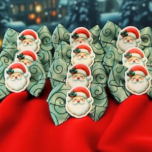 May include: A selection of Christmas hair bows. Each bow is light green with a swirl pattern and features a Santa Claus face. The bows are arranged on a red fabric background, with a blurred winter scene in the background.
