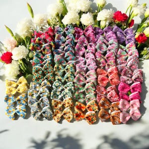 May include: A collection of 20 colourful floral print hair bows. The bows are arranged in a rainbow pattern, with each bow featuring a different floral design. The bows are made of fabric and have a white trim.